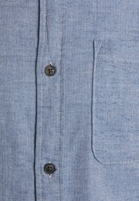 Tessuto di camicia con motivo a spina di pesce blu, due bottoni neri e una tasca quadrata cucita sul lato destro.