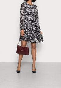 Schwarzes florales Kleid mit langen Ärmeln, gerafftem Saum und einer burgunderfarbenen Handtasche aus Kunst-Krokoleder. Model trägt schwarze spitze Pumps.