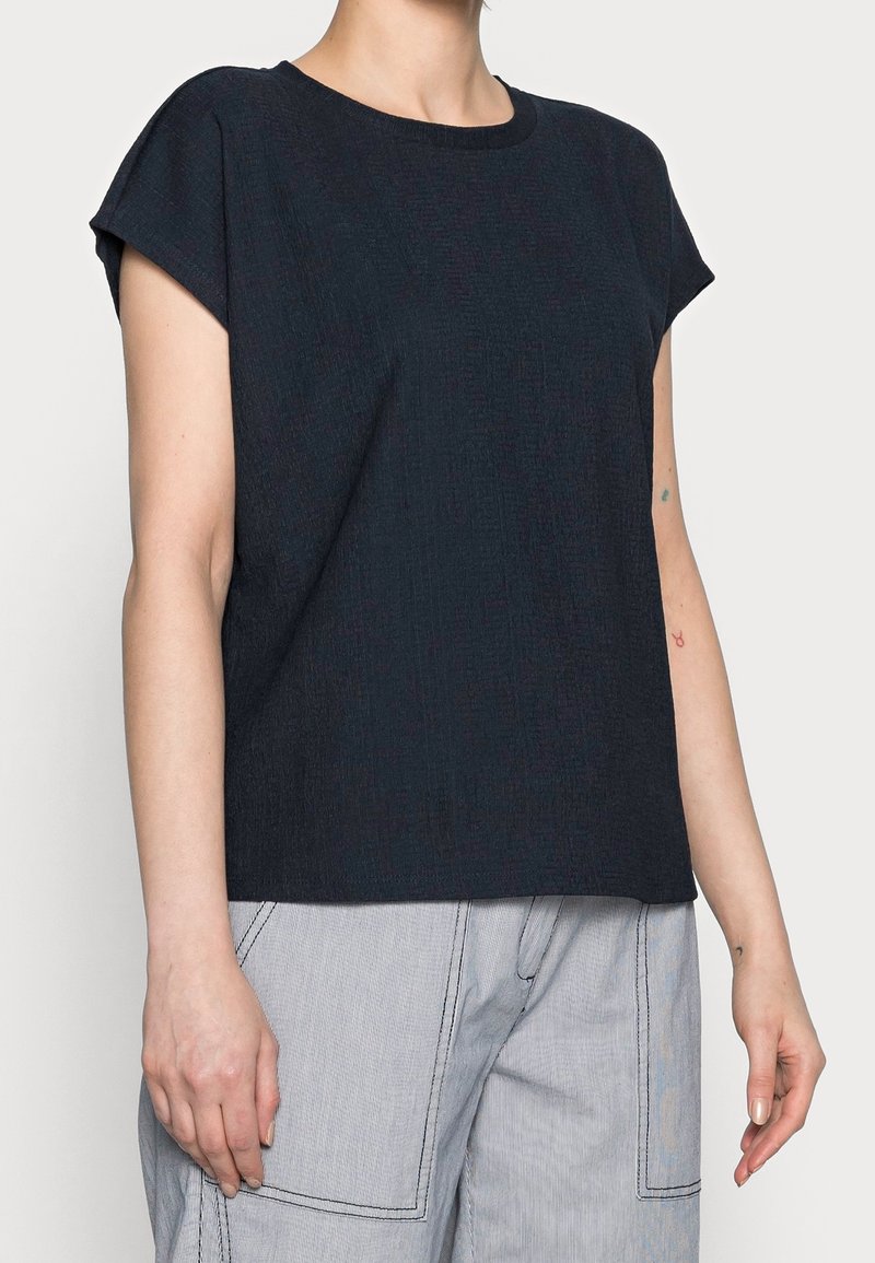 Námořnická modrá blůza s krátkým rukávem a kulatým výstřihem s jemným texturovaným vzorem. Vytvořeno s lehkými šedými pruhovanými kalhotami s širokými nohavicemi.