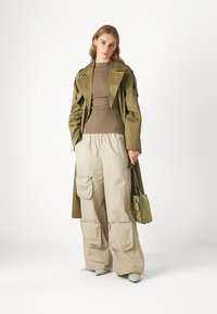 Olivenfarbener Trenchcoat über einem gerippten beigen Oberteil, kombiniert mit beigen Cargo-Hosen mit Seitentaschen und einer kleinen grünen Umhängetasche.
