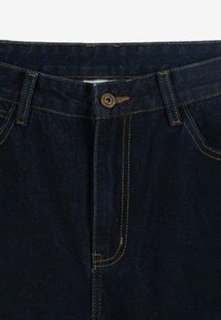 Granatowe dżinsy o klasycznym pięciopolowym wzorze, z brązowymi elementami metalowymi i złotymi szwami wzdłuż szwów. Gładka struktura.