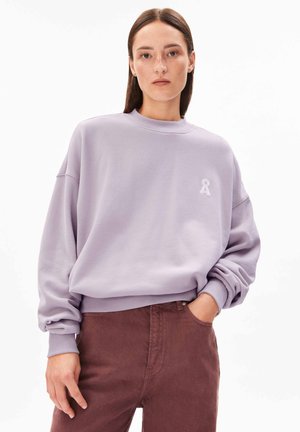 Woman with straight brown hair wearing a lavender sweatshirt with a small white emblem and maroon pants, hand in pocket, neutral background.