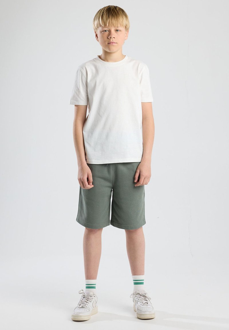 T-shirt bianca a maniche corte, pantaloni corti verdi, sneakers bianche con dettagli grigi e calze a righe. Design semplice con una vestibilità rilassata.