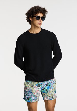 Black knit sweater with a round neckline paired with colorful, floral-patterned shorts. Model wears dark sunglasses.