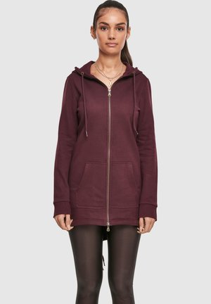 Femme portant un long sweat à capuche bordeaux zippé avec des poches avant et des leggings noirs, se tenant devant un fond clair.