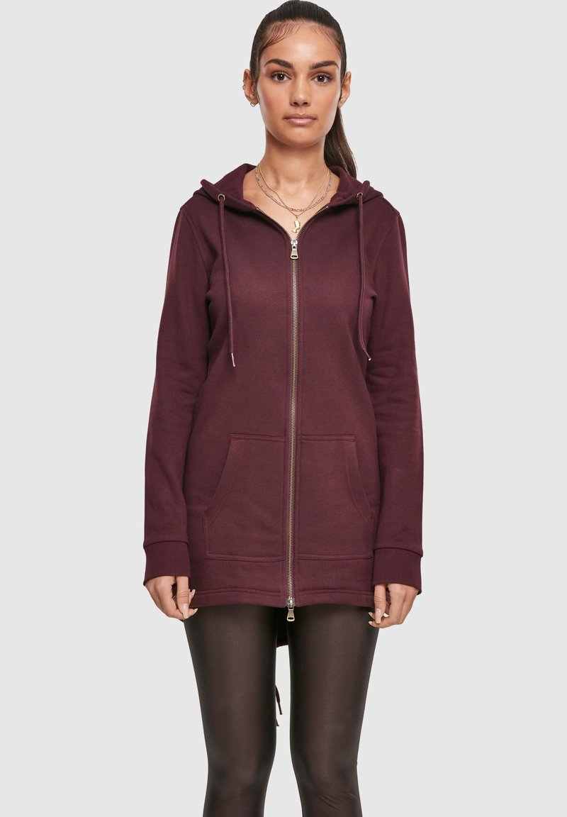 Žena u dugom burgundskom zip-up dukserici s prednjim džepovima i crnim tajicama, stoji na jednostavnoj svijetloj pozadini.