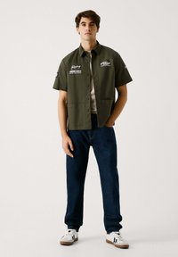 Camisa de manga corta verde oliva con detalles de logo, cierre de botones y dos bolsillos, combinada con jeans azul oscuro y zapatillas blancas.