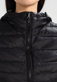 Czarna kurtka puchowa z wysokim kołnierzem, przednim suwakiem i regulowanymi sznurkami w kapturze. Posiada pikowaną wyściółkę dla lepszej izolacji cieplnej.