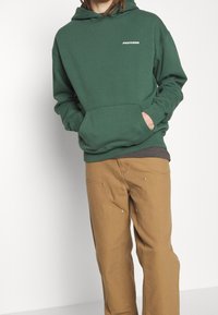 Mörkgrön hoodie med en framficka och en liten vit logotyp "PEGADOR" på vänster bröst, kombinerad med bruna byxor och en svart tröja under.