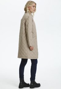 Manteau long beige matelassé avec col, poches latérales et accents de boutons ; porté sur un jean skinny noir et des bottines noires.