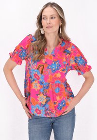 Blomstret bluse i lys pink med blå, orange og sorte accenter. Har korte, rynkede ærmer, en V-hals og en afslappet pasform.