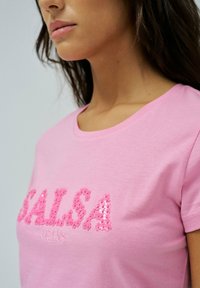 La camiseta de algodón rosa presenta la palabra "SALSA" en lentejuelas rosas en relieve, con un cuello redondo y mangas cortas. Textura suave, diseño casual.