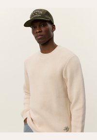 Beige Strickpullover mit rundem Ausschnitt, der ein strukturiertes Muster und einen Logoakzent am Saum aufweist. Dunkelgrüner Cordhut mit gesticktem Text.