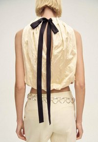 Back view of a sleeveless, cream-colored top with a keyhole opening, tied with a black satin ribbon at the neck; paired with beige pants and metallic belt.