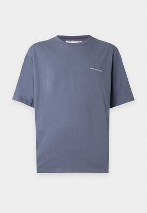 Homme debout portant un T-shirt bleu à manches courtes, un jean bleu clair et des chaussures marron à lacets sur fond blanc uni.