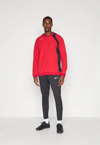 Férfi áll, piros és fekete Nike kapucnis pulóverben, fekete Nike melegítőnadrágban és fekete sportcipőben, egy sima világos háttér előtt.