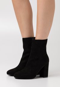 Bottes noires en daim jusqu'à la cheville avec une texture lisse, un bout pointu et un talon épais. Détail de fermeture éclair sur le côté intérieur pour un enfilage facile.
