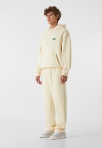 Sudadera crema y joggers a juego, hechos de una tela suave, con un bolsillo frontal y un detalle del logo verde en el pecho. Zapatillas visibles.