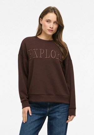 Frau trägt einen braunen Pullover mit dem Schriftzug "EXPLORE" und blaue Jeans, steht vor einem weißen Hintergrund und schaut leicht zur Seite.