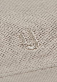 Világos bézs szövet, textúrált kötött mintával; jellemzi egy hangsúlyos, puha, fémpénz színű hímzett logó.