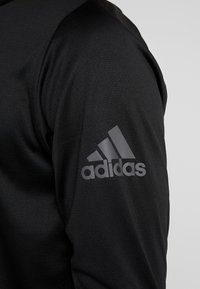 Camiseta atlética negra con tejido texturizado y un diseño ajustado. El hombro izquierdo presenta un logo de Adidas gris con tres rayas.