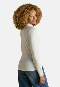 Langarm-Creme-Oberteil mit einem Rüschkragen, figurbetonter Silhouette und Knopfakzenten an den Ärmeln; kombiniert mit lässigen blauen Jeans.