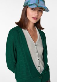 Grüner strukturiert Cardigan über einem hellgrauen Oberteil mit Knöpfen. Mütze mit blauen und grünen karierter Gestaltung. Kurzes, glattes Haar.
