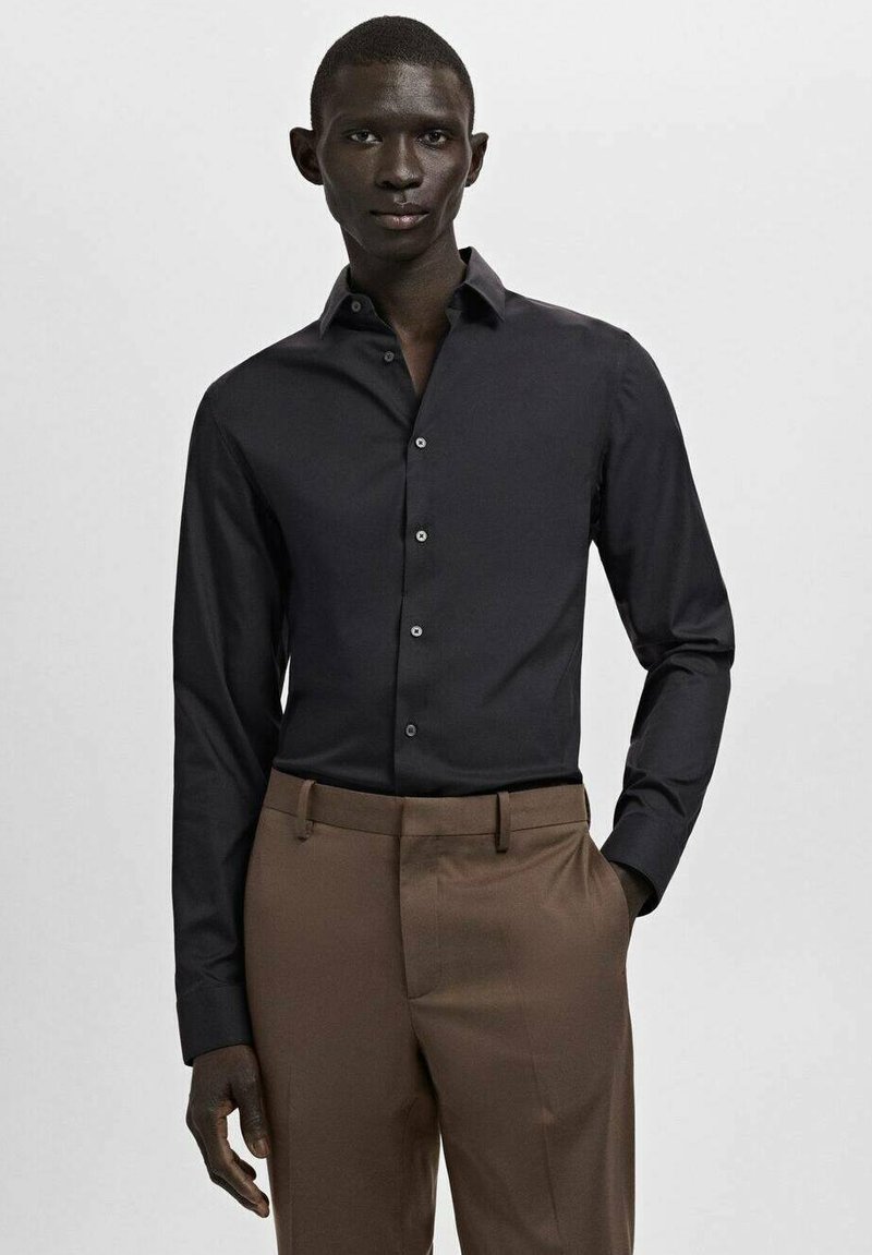 Chemise noire à boutons avec col pointu et manches longues, assortie à un pantalon marron clair. La chemise présente une texture lisse et une coupe ajustée.