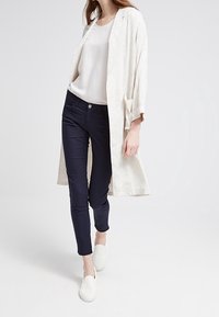Lätt, krämfärgad kimono-jacka över en vit topp, kombinerad med mörkblå slim-fit jeans och vita slip-on skor. Slät textur, avslappnad passform.