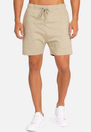 Mann trägt beige Shorts mit Kordelzug und weiße Turnschuhe, steht vor einem einfarbigen hellen Hintergrund.