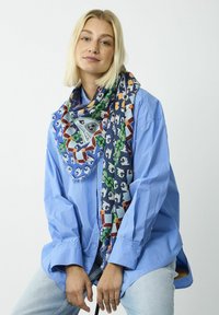 Écharpe multicolore avec des motifs géométriques et des bords frangés, portée sur une chemise oversized bleu clair ; le tissu semble léger et texturé.