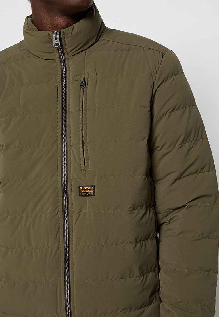 Hombre con chaqueta de acolchado en verde oliva, con cuello alto, cierre frontal, bolsillo vertical en el pecho y parche de logo de G-Star Originals.
