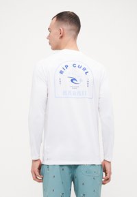 Langermet hvit skjorte med blå grafikk som viser "RIP CURL", en bølge, og "HAWAII" på baksiden. Glatt stoff, avslappet design.