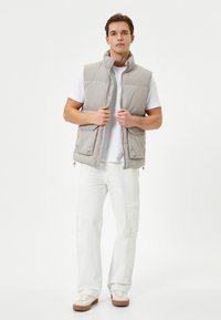Chaleco acolchado gris claro con un cuello alto, cierre frontal y dos bolsillos laterales, llevado sobre una camisa blanca y combinado con pantalones cargo blancos.