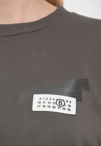 Primer plano de una sudadera gris que muestra una etiqueta numérica blanca con los números del 0 al 23 y el número 6 encerrado en un círculo, sujeto con cinta gris.