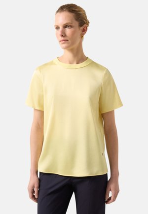 Femme portant une chemise légère jaune pâle à manches courtes et un pantalon sombre, debout avec une expression neutre devant un fond uni.