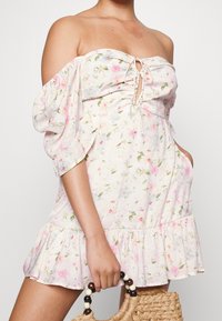Vestido floral con hombros al aire, mangas cortas abullonadas, tejido ligero, dobladillo con volantes y frente con cordón ajustable. Presenta un suave patrón en tonos rosa y verde.