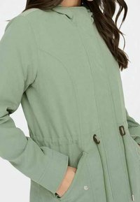 Světlý zelený kabát s kapucí, který má stáhnutý pas, boční kapsy a kontrastní hnědé detaily. Hladká tkanina s jemnou texturou.