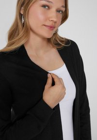 Schwarze Strickjacke mit V-Ausschnitt, über einem weißen Tanktop getragen, mit weichem Stoff und langen Ärmeln. Sichtbare silberne Creolen.