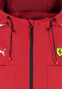 Czerwona kurtka z kapturem, wykonana z teksturowanej tkaniny, z czarnym zamkiem błyskawicznym, regulowanymi sznurkami oraz żółtym logo Ferrari na lewej piersi.