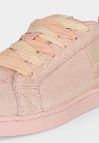 Gros plan d'une basket en daim rose avec des larges lacets couleur pêche pastel et crème et une semelle en caoutchouc texturée.