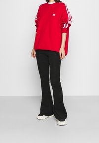 Röd överdimensionerad sweatshirt med vita ränder på ärmarna, vit logotyp på bröstet, tillsammans med svarta ribbade utsvängda byxor och grova sneakers.