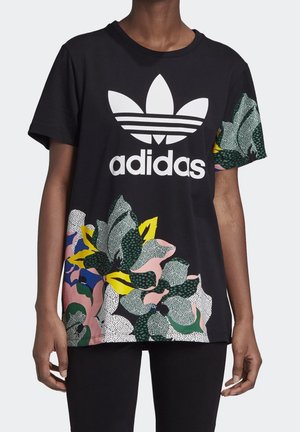 Czarny t-shirt Adidas z dużym białym logo na piersi oraz kolorowym, abstrakcyjnym wzorem liści na dolnej części przodu i prawym rękawie, noszony przez osobę.