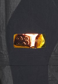 Svart tyg med en blank guldrektangulär badge som visar "23" och märkdetails. Strukturerad yta och kontrasterande rutmönster.