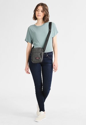 Sac à bandoulière gris avec deux poches zippées, fabriqué en tissu texturé. Le modèle porte un haut teal, un jean foncé et des baskets claires.
