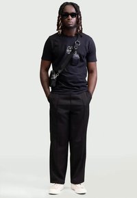 Black graphic t-shirt with white text, black trousers, sunglasses, crossbody bag, and white sneakers. Casual urban outfit.