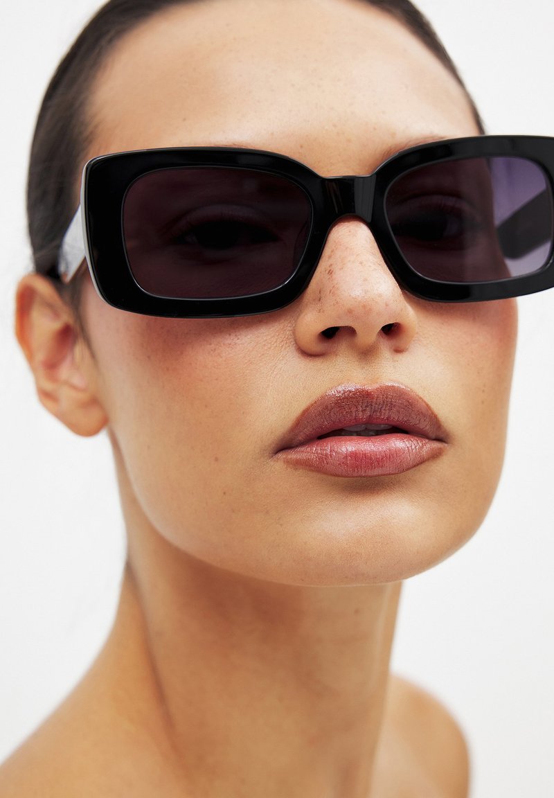 Črne pravokotne sončna očala s temnimi lečami, debelimi sijajnimi okvirji in svetlimi stranskimi akcenti. Gladka koža in naravna barva ustnic.