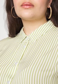 Camisa de cuello a rayas verdes y blancas con cierre de botones; textura suave y tela ligera. Grandes pendientes en forma de aro visibles.