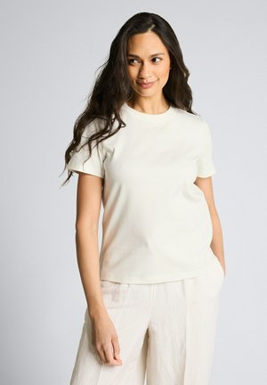 Kobieta o długich ciemnych włosach, ubrana w białą koszulkę z krótkim rękawem i białe spodnie w paski, stoi z jedną ręką w kieszeni, patrząc na bok.