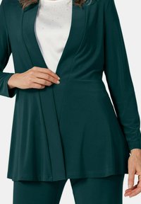 Diep groenblauwe blazer met lange mouwen en een vloeiende lijn. Zachte stof, open voorkant en subtiele draping. Draagt over een witte top met versieringen.